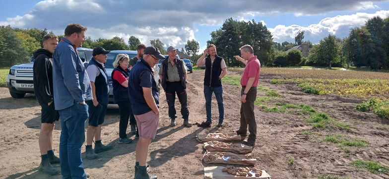 A group of people on a farm, listening to a man speak. Potatoes are displayed on the ground.