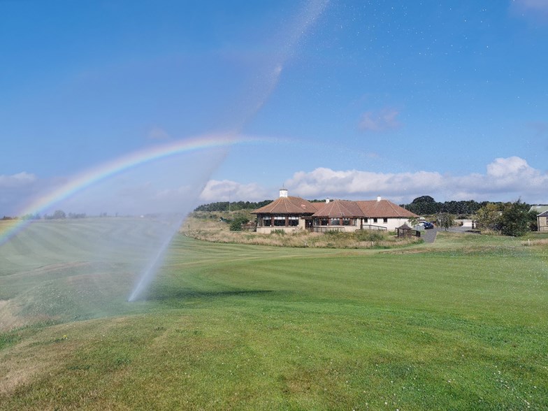 A golf course with a sprinkler spraying water into the sky, forming a rainbow.