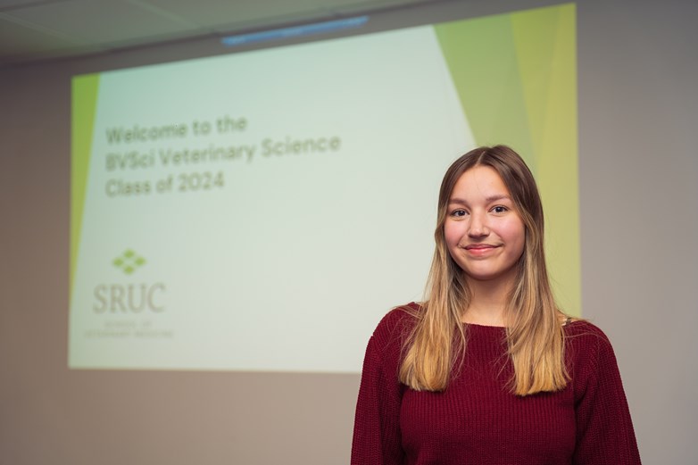 A young woman stands in front of a PowerPoint presentation. On the screen it says 'Welcome to the BVSci Veterinary Science Class of 2024' along with the SRUC logo.