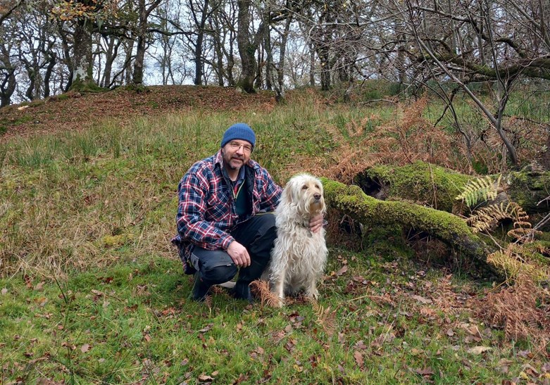 A man crouching in the woods with his dog.